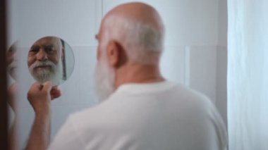 Shooting over shoulder of blurred old man admiring reflection in hand mirror touching long white beard. Smiling happy Caucasian male retiree at home in the morning. Slow motion