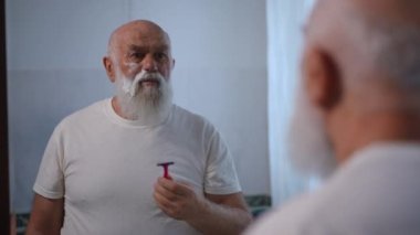 Reflection in mirror of senior Caucasian man shaving stubble on cheek with razor. Concentrated confident old retiree styling white beard at home in bathroom. Hygiene and lifestyle concept