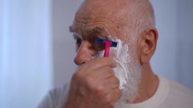 Headshot of gray-haired old Caucasian man shaving with razor in slow motion indoors. Focused senior retiree styling white beard in bathroom at home indoors. Lifestyle and hygiene concept