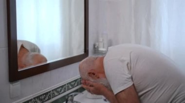 Confident old man washing off shaving foam leftovers from face looking in bathroom mirror. Concentrated senior Caucasian retiree styling white long beard in the morning at home indoors