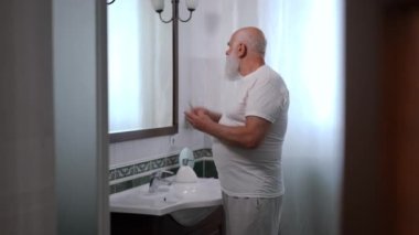 Confident senior bearded man combing white long beard gesturing thumb up to reflection in mirror leaving bathroom. Portrait of handsome positive Caucasian retiree at home in the morning