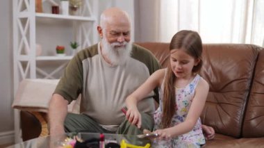 Charming cute Caucasian granddaughter applying face powder on face of smiling positive grandfather sitting on couch in living room. Happy relaxed pretty girl and handsome man enjoying weekend at home