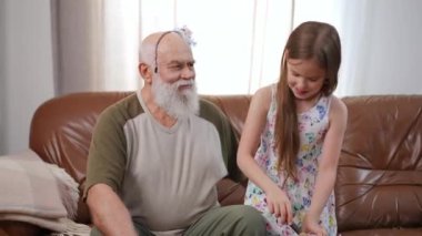 Laughing senior man in hair hoop sitting on couch as girl applying face powder on forehead with brush. Happy carefree Caucasian grandfather having fun playing with granddaughter at home in living room