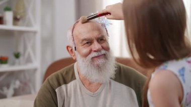 Portrait of laughing old Caucasian grandfather in hair hoop admiring granddaughter applying face powder on forehead. Happy relaxed senior man enjoying weekend with charming girl at home indoors