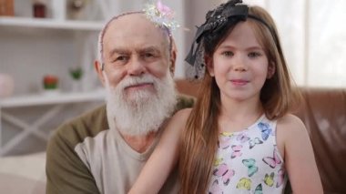 Happy granddaughter and grandfather in hair hoops posing at home in living room. Caucasian senior bearded man and cute charming girl looking at camera smiling enjoying weekend leisure indoors
