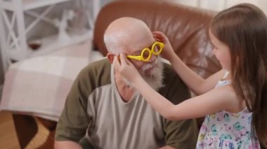 Positive cheerful girl putting on toy yellow glasses on face of senior man smiling. Confident positive Caucasian grandfather playing with granddaughter on weekend at home indoors hugging child