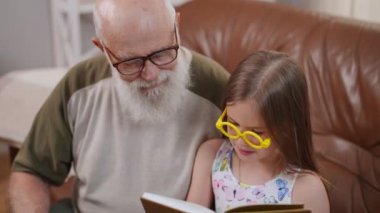 High angle view portrait of concentrated Caucasian old grandfather and cute focused granddaughter reading book in slow motion sitting at home. Relaxed confident man and girl enjoying hobby