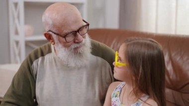 Senior grandfather in eyeglasses and charming granddaughter in yellow toy glasses looking at each other turning looking at camera smiling. Positive Caucasian old man and girl posing in slow motion