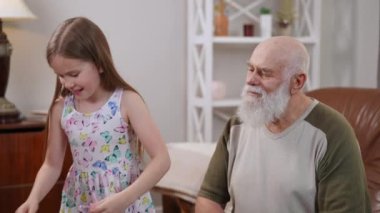 Bearded positive senior Caucasian man talking with pretty girl sticking toy gems on retiree forehead passing hand mirror laughing. Carefree grandfather and granddaughter enjoying weekend having fun