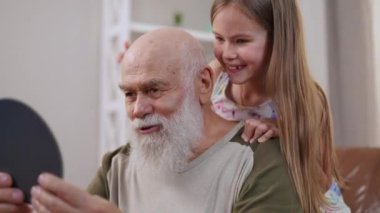 Positive senior man laughing talking looking at hand mirror with excited girl placing toy gems on forehead of retiree. Happy carefree Caucasian grandfather playing with granddaughter enjoying leisure