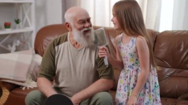 Funny Caucasian granddaughter and grandfather singing in hairbrush enjoying leisure on weekend in living room. Relaxed happy cute girl and senior man having fun at home indoors