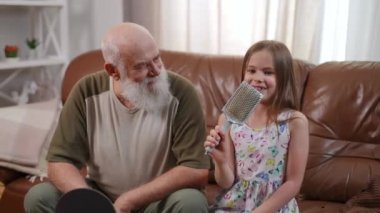 Hyperactive pretty girl singing in hairbrush sitting with smiling old bearded man on sofa in living room. Positive joyful Caucasian granddaughter enjoying hobby with grandfather at home