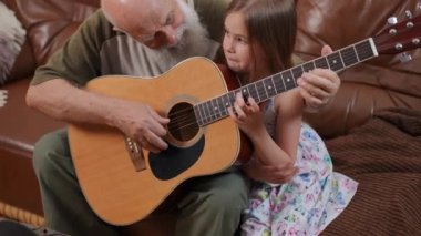Smiling satisfied senior grandfather teaching playing guitar sitting with curious granddaughter on sofa in living room. Portrait of happy confident Caucasian old man and charming girl enjoying hobby