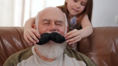 Front view close-up of positive senior bearded man with blurred girl placing toy black mustache on face of retiree. Relaxed Caucasian grandfather on weekend with playful granddaughter. Slow motion