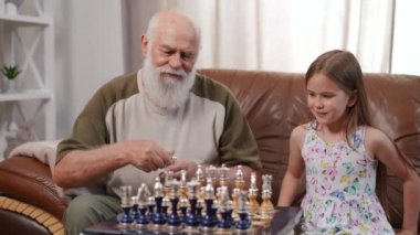 Cheerful curious girl choosing chess color clapping playing with old bearded man at home indoors. Portrait of joyful Caucasian granddaughter enjoying hobby with grandfather in living room