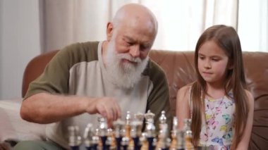 Intelligent senior grandfather talking with granddaughter teaching child playing chess. Concentrated curious Caucasian cute girl listening to smart bearded old man sitting on couch in living room