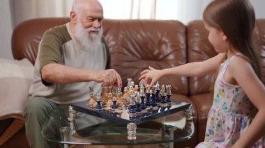 Chessboard on table with senior Caucasian bearded man and curios pretty girl thinking choosing piece. Confident intelligent grandfather and granddaughter playing game at home on weekend