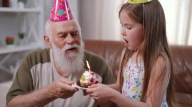 Senior man and little girl blowing out burning candle on cupcake in slow motion. Smiling happy Caucasian grandfather and granddaughter celebrating birthday at home indoors