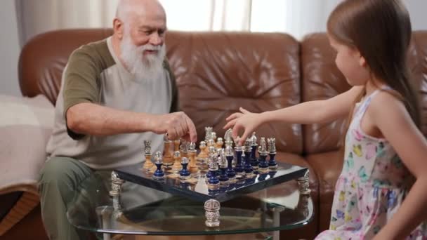 上級白人ひげを生やした男と骨董品でテーブルの上のチェス盤かなり女の子は作品を選択することを考えています 自信を持ってインテリジェント祖父と孫娘は週末に自宅でゲームをプレイ — ストック動画