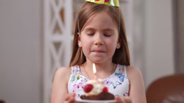 Close-up portrait of pretty charming Caucasian girl making wish and blowing out burning candle on birthday cupcake in slow motion. Happy cute child in party hat looking at camera smiling celebrating