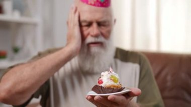 Close-up sweet delicious cupcake in senior male hand with blurred old man talking at background. Sad Caucasian bearded retiree celebrating birthday alone at home. Aging and sadness concept