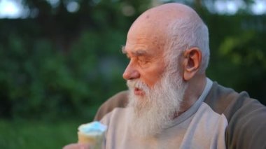 Close-up happy senior bearded man enjoying delicious ice cream looking away talking. Relaxed Caucasian old retiree eating dessert in summer park at sunset outdoors. Slow motion