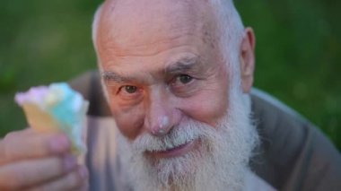 High angle view close-up of joyful old man with icing on nose showing ice cream eating delicious dessert in slow motion. Happy carefree Caucasian retiree enjoying tasty food posing in summer park