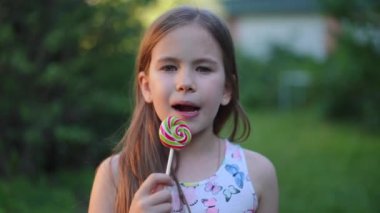 Close-up front view of pretty brunette Caucasian girl with brown eyes looking at camera licking multicolored lollipop in slow motion. Happy satisfied child posing in summer park enjoying tasty dessert