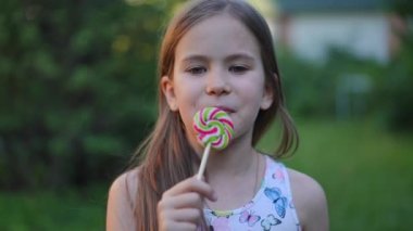 Portrait of happy satisfied cute slim girl posing eating delicious lollipop standing in summer park. Pretty Caucasian child looking at camera and looking away enjoying tasty colorful dessert