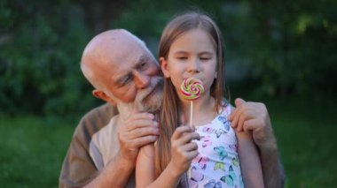 Live camera zoom in to smiling pretty Caucasian girl posing with senior bearded man and colorful lollipop in summer park. Happy confident granddaughter enjoying picnic with grandfather outdoors