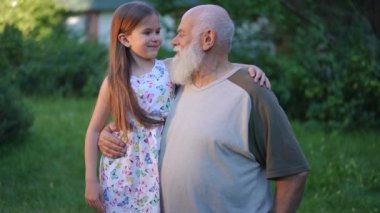 Zoom in portrait of hugging happy Caucasian senior grandfather and granddaughter smiling and looking at camera. Positive confident old bearded man and charming cute girl posing in slow motion in park