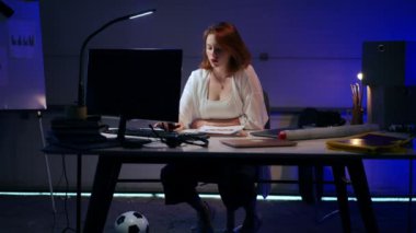 Wide shot portrait of concentrated freelancer analyst entering data in PC analyzing paperwork statistics. Redhead beautiful Caucasian young woman working online sitting in garage thinking