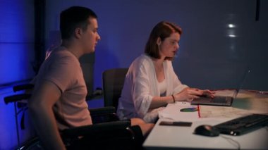 Side view positive focused young confident woman surfing Internet on laptop talking with man in wheelchair in garage. Concentrated Caucasian startupers analyzing e-market planning business idea