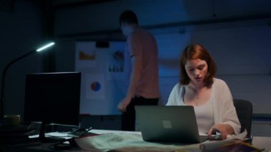 Portrait of confident focused woman messaging online on laptop as blurred man analyzing paperwork standing at whiteboard at background. Beautiful startuper planning e-business with colleague in garage