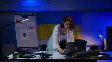 Young charming intelligent woman analyzing paperwork typing on laptop keyboard standing at table smiling. Portrait of smart Caucasian confident freelancer working online in garage indoors