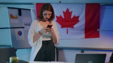 Young redhead woman messaging in smartphone app standing in garage with Canadian flag smiling. Absorbed millennial freelancer planning startup idea scrolling touchscreen