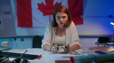 Front view absorbed freelancer gamer looking at camera playing video game with joystick losing leaning on chair. Portrait of redhead concentrated Caucasian millennial woman gaming indoors on break