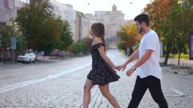 Side view portrait of confident beautiful woman crossing road holding hand of handsome man. Wide tracking shot of loving millennial Caucasian boyfriend and girlfriend walking on urban city street