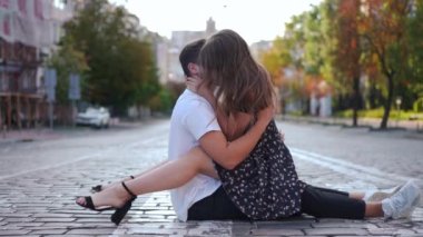 Side view happy young couple sitting on urban road hugging and kissing as woman turning looking at camera smiling. Satisfied confident beautiful Caucasian girlfriend posing with loving boyfriend