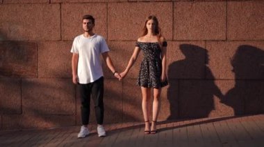 Wide shot front view of confident beautiful woman looking at camera holding hands with man looking away. Happy young Caucasian couple posing at background of urban city wall on sunny street