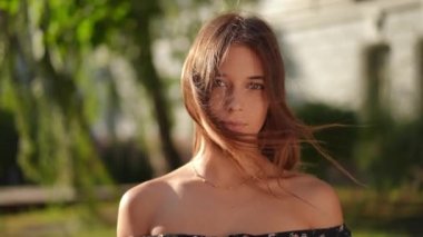 Close-up front view of smiling gorgeous young woman looking at camera caressing long brunette hair in slow motion. Positive confident Caucasian millennial slim lady posing on city street at sunset