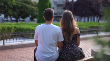Back view happy relaxed couple sitting on bench on city street as woman putting head on man shoulder in slow motion. Medium shot of unrecognizable young man and woman dating outdoors