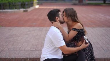 Side view happy woman kissing man in slow motion sitting on curb in city. Medium shot portrait of loving Caucasian talented photographer dating with girlfriend outdoors hugging