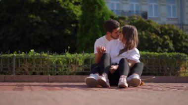 Wide shot portrait of loving happy relaxed couple kissing sitting on the right on skateboard outdoors. Relaxed confident Caucasian boyfriend and girlfriend dating on city street hugging cuddling