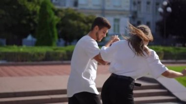 Side view young woman learning riding skateboard with man helping holding hand walking aside. Live camera panning as confident Caucasian girlfriend and boyfriend enjoying hobby dating on city street
