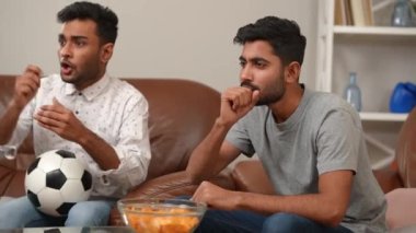 İki heyecanlı arkadaş, futbol topu ve cipslerle kanepede oturup el kol hareketi yapıyor. Pozitif genç Orta Doğulu erkekler TV 'de futbol maçı izleyip hafta sonunun keyfini çıkarıyorlar.
