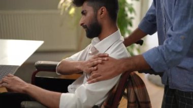 Dizüstü bilgisayarı olan tekerlekli sandalyedeki stresli genç bir adam ve tanınmayan bir arkadaşı omzuna dokunuyor. Minnettar Orta Doğulu adam içeride avuç içi ve mesajlaşmalara dokunuyor.
