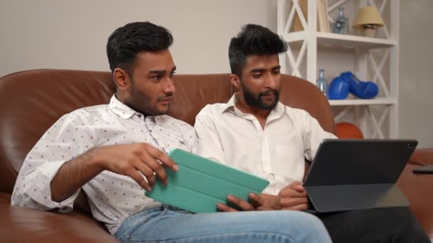 2人の思慮深い濃縮された中東の男性が居心地の良いリビングルームで快適なソファに座っているデジタルタブレットでインターネットサーフィンを話しています 自宅でビジネスを計画するインテリジェントな自信のある友人 — ストック動画