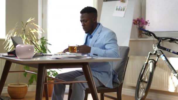忙しいアフリカ系アメリカ人の若者が自宅のオフィスでパンケーキを食べてお茶を飲んでテーブルに座っている ラップトップ上の電子メールをチェック午前中に集中深刻な千年のスタートアップ企画ビジネス — ストック動画