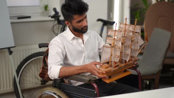 坐在室内轮椅上的带木船模型的中东工匠的画像 英俊而自信的年轻人在总公司欣赏手工制作的艺术品 — 图库视频影像